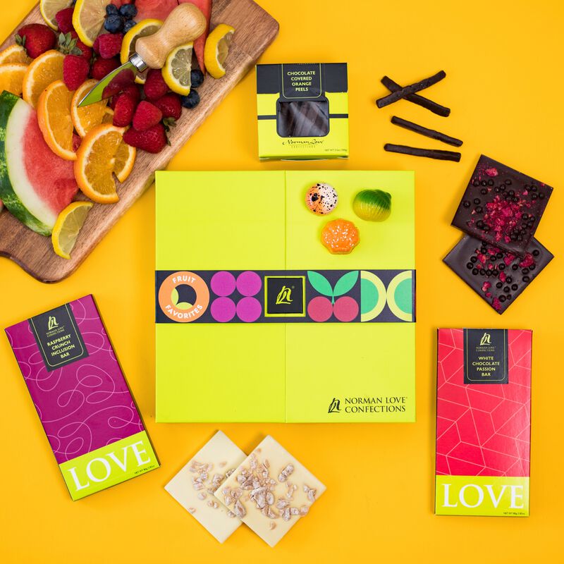 LIme green truffle box with fruit flavored candies on top. Plate of fruit, square pieces of dark chocolate raspberry inclusion bar and white chocolate passionfruit inclusion bar and chocolate covered candied orange peels shown on orange background. 